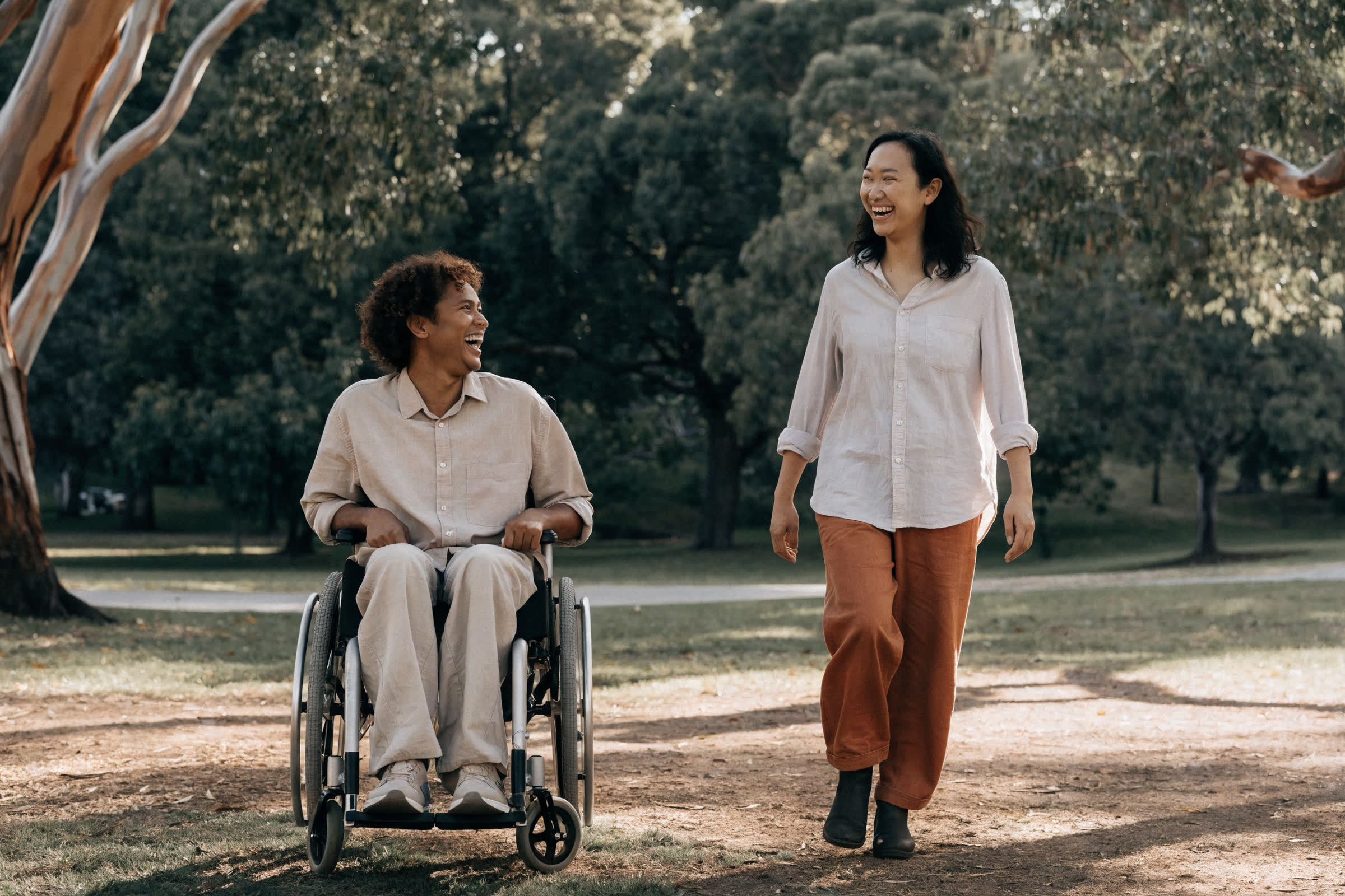 An adult wheelchair user and a support worker sharing a relaxed moment in a leafy Sydney park